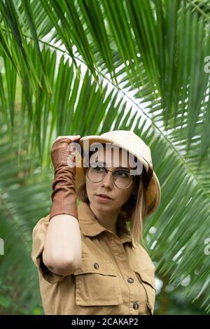 Donna botanica vestita in stile safari in serra. Naturalista in abiti cachi, guanti di lievito nella foresta pluviale circondati da palme. Giungla turistico Foto Stock