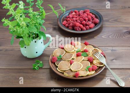 Pancake cereali sulla forchetta. Lamponi freschi maturi rossi in un piatto marrone e un mazzo di Melissa in una tazza su un tavolo di legno scuro. Deliziosa Brea estiva Foto Stock