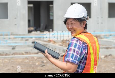 Ritratto di donna asiatica tecnico delle costruzioni lavoratore con il casco in testa usando tablet mentre si sta in piedi sul sito in costruzione. cantiere posto sul retro Foto Stock