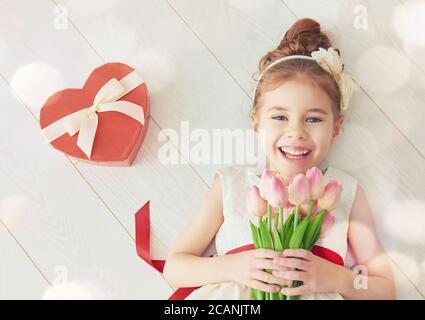 Bambina dolce con cuore rosso e bouquet di tulipani. Felice bambina con regalo di San Valentino sdraiato su pavimento di legno bianco. Matrimonio, concetto di San Valentino Foto Stock