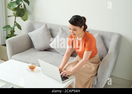 Giovane donna che si rilassa a casa sul divano, sta facendo un tè e sta usando un computer portatile Foto Stock