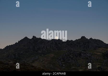 La luna quasi piena si alza lentamente dietro le pietre sulle cime di montagna in un momento crepuscolo, lontano in lontananza Foto Stock