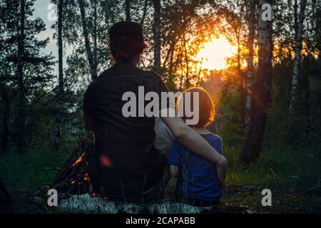 Padre e figlio sono seduti su un log accanto al fuoco e guardando il tramonto con le spalle. Sfondo della foresta. Foto Stock