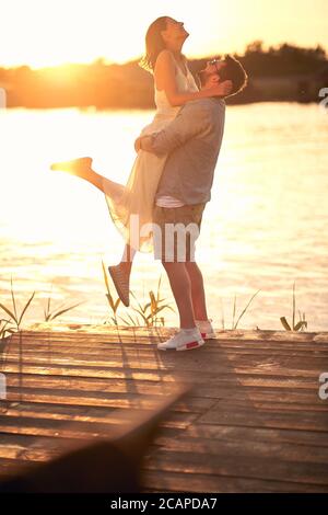 ragazzo caucasico ardito abbracciare, sollevando la sua fidanzata al tramonto dal fiume Foto Stock