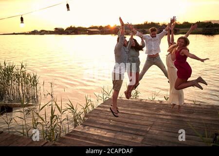 gruppo di giovani amici adulti che saltano sulla piattaforma di legno vicino al fiume, divertendosi Foto Stock