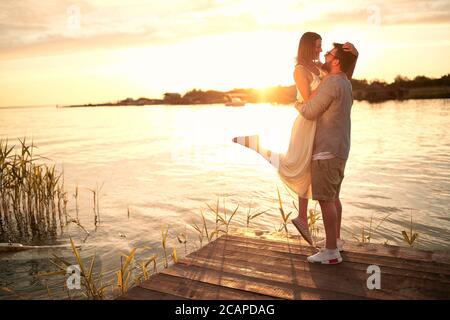 ragazzo caucasico che tiene in mano la sua ragazza al tramonto sul fiume Foto Stock