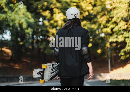 Il ragazzo con la tavola cammina per la strada. Longboarding al tramonto. Foto Stock