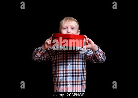 Piccolo ragazzo che tiene una scatola a forma di cuore rosso. Isolare su sfondo nero. Foto Stock