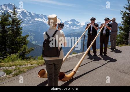 Schynige Platte, Oberland Bernese, Svizzera - 1 agosto 2019 : 2 giocatori di Alpenhorn in abito svizzero tradizionale girato da turista femminile su cellulare ph Foto Stock