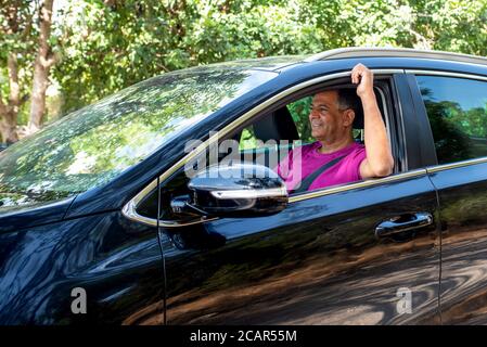 Vista di profilo di un uomo anziano felice nella sua metà degli anni '60 al volante di una macchina. Uomo che guida auto in foresta. Foto Stock