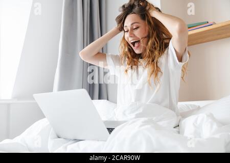 Ritratto di donna zenzero eccitato esprimendo sorpresa e utilizzando il laptop mentre si siede a letto dopo il sonno Foto Stock