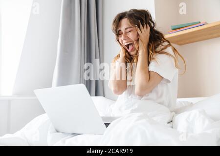 Ritratto di donna zenzero eccitato esprimendo sorpresa e utilizzando il laptop mentre si siede a letto dopo il sonno Foto Stock