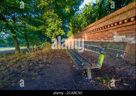 Una vecchia e rotta panchina a Harvington Park, Beckenham, Kent. La panchina è al tramonto con un muro di mattoni rossi e alberi dietro. Foto Stock