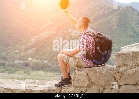 Giovane uomo con zaino si siede su parete di pietra con rialzato cappello sullo sfondo di montagne verdi Foto Stock