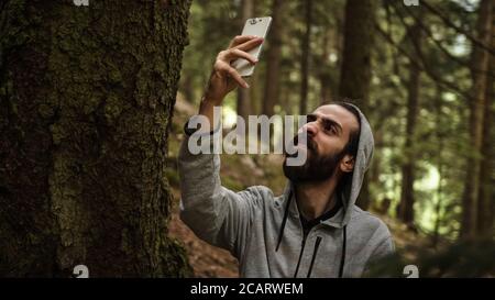 L'adolescente hipster che testava nella foresta con un telefono in mano rompe il segnale, stressava il giovane uomo con problemi di segnale Foto Stock