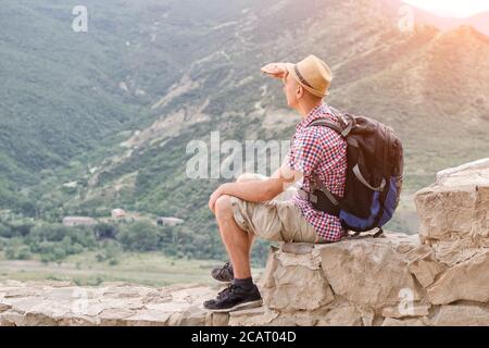 Un giovane uomo in un cappello con uno zaino si siede sopra un muro di pietra contro uno sfondo di montagne verdi e. guarda in lontananza Foto Stock