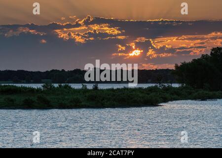 Il sole sbirciando attraverso le nuvole del mattino presto su un lago con una penisola ricoperta di alberi che divide le acque in quello che sembra due laghi. Foto Stock