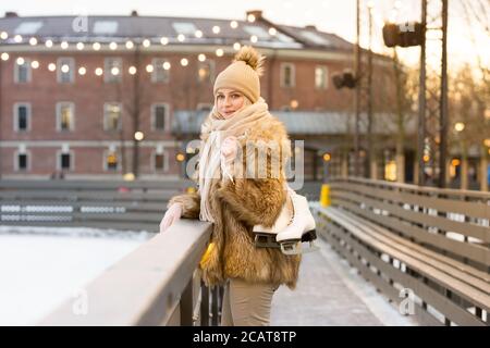 Giovane donna europea allegra alla pista di pattinaggio su ghiaccio che tiene pattini bianchi e sorridente, all'aperto in gelido giorno d'inverno.stile di vita sano, sport e tempo libero Foto Stock