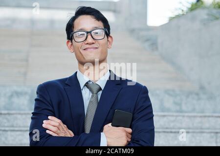 Felice orgoglioso giovane dirigente d'affari asiatico in glase con smartphone e sorridendo alla macchina fotografica Foto Stock