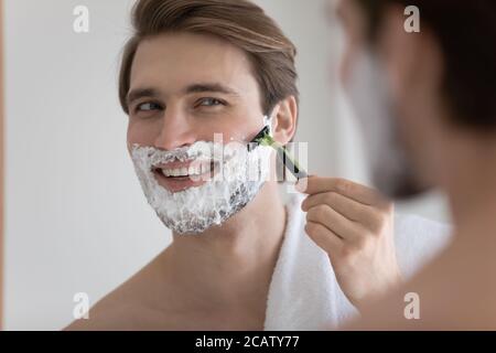 Primo piano riflessione specchio sorridente uomo che si rade, utilizzando il rasoio Foto Stock