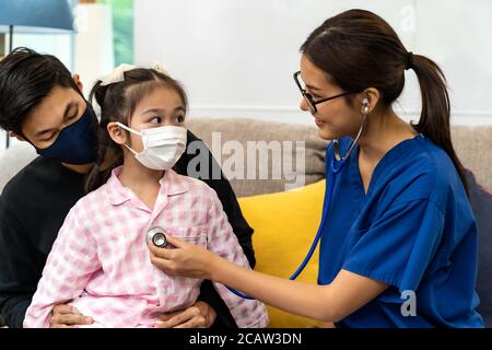 Il medico femminile asiatico usa stetoscopio per ascoltare il battito cardiaco della bambina si siede con suo padre in soggiorno mentre il medico visita a casa. Healt domestico Foto Stock