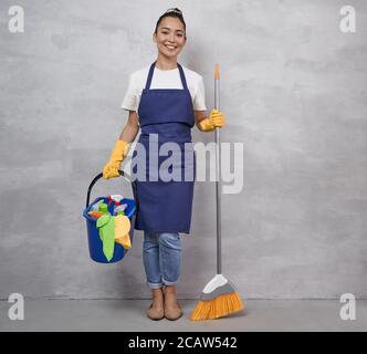 Donna di pulizia felice in uniforme e giallo guanti di gomma che reggono scopa e secchio di plastica con stracci e diversi prodotti per la pulizia mentre si trova contro parete grigia. Servizi di pulizia, pulizie Foto Stock