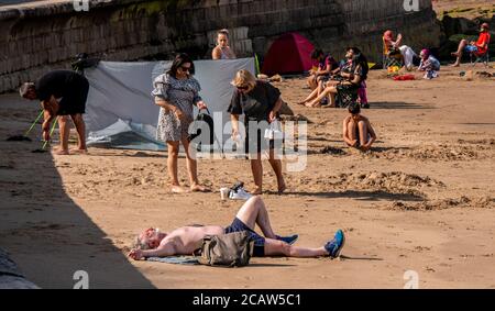 Un uomo si addormenta sulla spiaggia di Scarborough con le braccia distese e gli altri bagnanti sullo sfondo. Foto Stock