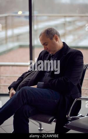 Stazione ferroviaria. Uomo maturo in abiti da lavoro sta dormendo, seduto in sala d'attesa e in attesa di treno. La sera è buia. Foto Stock