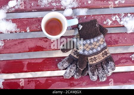 Tazza di tè e guanti in maglia da donna su una panca in inverno, la neve sotto. Vista dall'alto Foto Stock