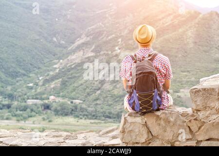 Un giovane uomo in un cappello con uno zaino si siede su un muro di pietra contro uno sfondo di montagne verdi. Vista posteriore Foto Stock