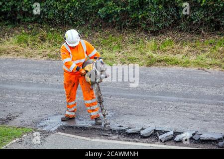 Lavorare la donna con il martello di sollevamento che esegue riparazioni stradali e riparazione dei danni superficie stradale irregolare e fori di pentola con asfalto in a. Cheshire Country Lane Road Foto Stock