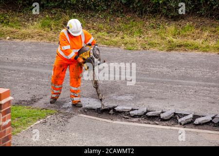 Lavorare la donna con il martello di sollevamento che esegue riparazioni stradali e riparazione dei danni superficie stradale irregolare e fori di pentola con asfalto in a. Cheshire Country Lane Road Foto Stock