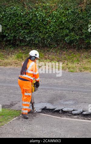 Lavorare la donna con il martello di sollevamento che esegue riparazioni stradali e riparazione dei danni superficie stradale irregolare e fori di pentola con asfalto in a. Cheshire Country Lane Road Foto Stock