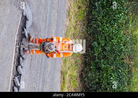 Lavorare la donna con il martello di sollevamento che esegue riparazioni stradali e riparazione dei danni superficie stradale irregolare e fori di pentola con asfalto in a. Cheshire Country Lane Road Foto Stock