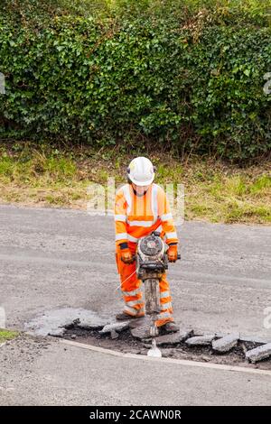 Lavorare la donna con il martello di sollevamento che esegue riparazioni stradali e riparazione dei danni superficie stradale irregolare e fori di pentola con asfalto in a. Cheshire Country Lane Road Foto Stock