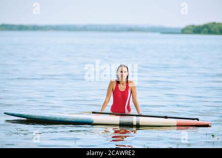 Felice giovane donna bruna in costume da bagno rosso in piedi in acqua con tavola da surf Foto Stock