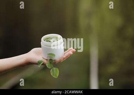 le mani della donna che tengono il vaso di gel cosmetico di aloe vicino a. foglie verdi e rami sullo sfondo della natura Foto Stock