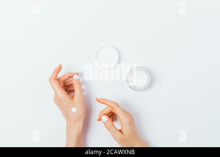 Belle mani di giovane donna con manicure applicare crema cosmetica su tavola bianca, composizione piatta. Foto Stock