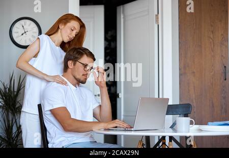 adorabile donna rosso sostegno marito lavorante, giovane donna attenta lo abbraccia, chiede come si sente, è stanco, cosa fare per lui Foto Stock