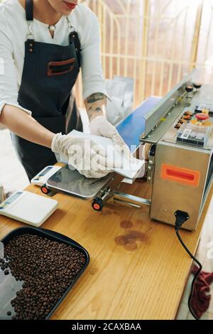 Lavoratrice di maestria femminile in grembiule e guanti in gomma sigillano le cuciture di pacchetti di caffè Foto Stock