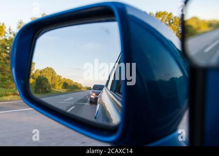 Vista posteriore nello specchietto laterale di una berlina blu con il riflesso di un crossover grigio che mostra un segnale di svolta di una manovra di sorpasso su un asp Foto Stock
