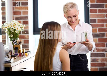 sorridente adulto make-up artista guardare arrossire prima di applicarlo sul viso, professionale visage master godere, ama il suo lavoro Foto Stock