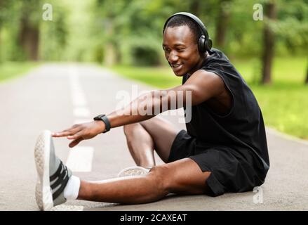 Sorridente sportivo nero seduto sul sentiero al parco, stretching Foto Stock