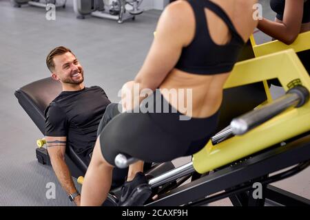 le persone atletiche si sostengono in palestra, fare esercizi insieme. uomo pompando gambe su attrezzature con peso, vista posteriore su donna seduta vicino, uomo smill Foto Stock