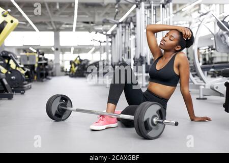 donna afro-fit nera stanca dopo esercizi sollevamento pesi, giovane donna siede accanto al barbell, esausta Foto Stock