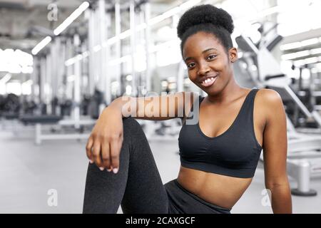ritratto di adorabile donna africana seduta in palestra, giovane donna in abbigliamento sportivo dopo esercizi di crossfit, posa, sorriso. concetto di stile di vita sano Foto Stock