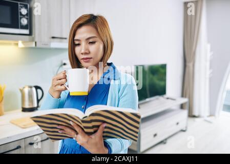 Bella giovane donna vietnamita godendo interessante libro e bere tazza di caffè forte Foto Stock
