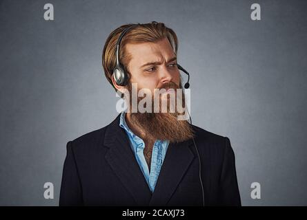 Uomo bello lavorare come un servizio di assistenza clienti, indossa la cuffia alle chiamate, guarda scontroso e annoiato, sconvolto e gravi barbuto hipster business man. Foto Stock
