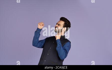 Concetto di successo. Eccitato ragazzo indiano sentirsi vittorioso e gesturing si su sfondo lilla Foto Stock