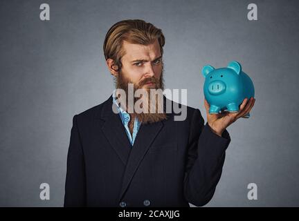 Finanza, denaro. Grave uomo detiene il salvadanaio. Uomo Barbuto detiene il salvadanaio pieno di denaro su sfondo grigio. Aziendali e finanziarie; il finanziamento della misura Foto Stock